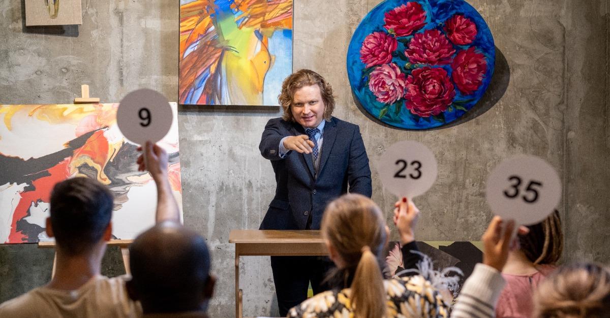 Mature male auctioneer in formalwear standing by tribune and pointing at one of people with auction paddles